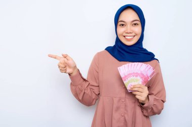Pembe gömlekli gülümseyen Asyalı Müslüman kadın elinde para banknotları ve beyaz arka planda izole edilmiş boş bir yeri işaret ediyor. İnsanlar dini yaşam tarzı konsepti