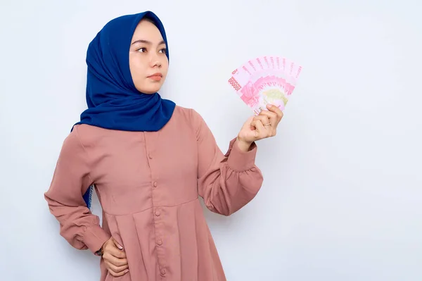 confused young Asian muslim woman in pink shirt  holding money banknotes and looking away at copy space isolated over white background. People religious lifestyle concept