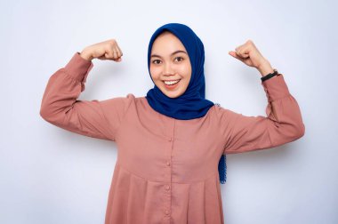 Pembe gömlekli, gülümseyen Asyalı Müslüman kadın kollarını kaldırıyor ve pazılarını gösteriyor. Beyazların arka planında izole edilmiş bir spor salonunda aldığı eğitimden sonra başarısını gösterdi. İnsanlar dini yaşam tarzı konsepti