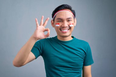 Smiling young Asian man showing ok sign celebrating Indonesia independence day isolated on gray background. Indonesian independence day celebration concept