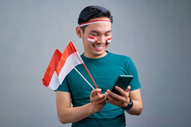 Smiling young Asian man holding mobile phone and indonesian flag and looking at camera isolated on grey background. indonesia independence day celebration concept
