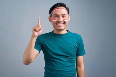 Cheerful young Asian man in casual t-shirt pointing finger up, having an idea or found solution isolated on grey background. indonesian independence day celebration concept