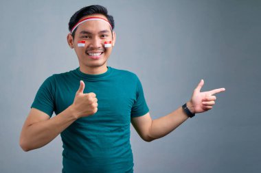 Smiling young Asian man in casual t-shirt  pointing finger aside at copy space, showing thumb up isolated on grey background. indonesian independence day celebration concept