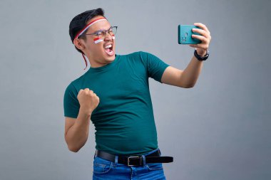 Excited young Asian man in casual t-shirt playing online game on mobile phone, celebrating victory isolated on grey background. indonesian independence day celebration concept