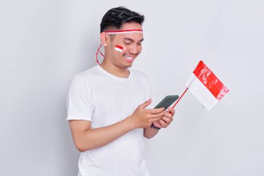 Cheerful young Asian man using mobile phone and celebrating Indonesia independence day on August 17 isolated on white background