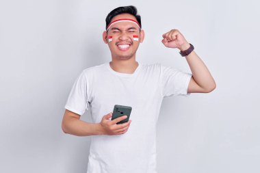 Excited and confident young Asian man holding mobile phone, clenched both fist to celebrating Indonesia independence day on August 17 isolated on white background