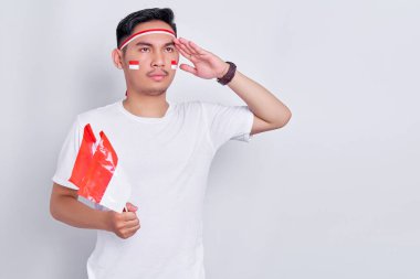Portrait of smiling young Asian man holding indonesian flag and showing respect gesture while celebrating indonesian independence day on 17 august isolated on white background
