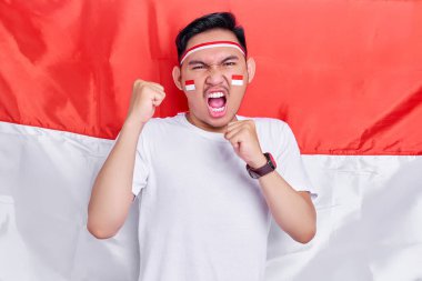 Young Asian man clenched both fist to celebrate Indonesia independence day on August 17, looking at camera with confident and excited expression isolated on indonesian flag background