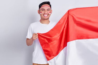 Smiling young Asian man holding indonesian flag and showing respect gesture while celebrating indonesian independence day on 17 august isolated on white background