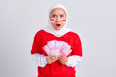 Amazed young Asian muslim woman in red white t-shirt holding cash money in Indonesian rupiah banknotes isolated on white background. Indonesian independence day on 17 august concept