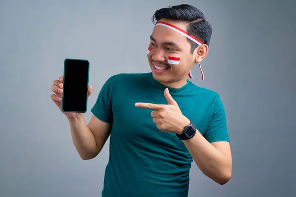 Cheerful young Asian man in casual t-shirt pointing fingers at blank screen mobile phone, recommending mobile app isolated on grey background. indonesian independence day celebration concept