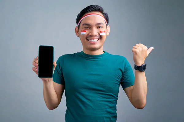 Smiling young Asian man showing mobile phone with blank screen, recommending mobile app, pointing thumb aside at copy space isolated on grey background. indonesian independence day celebration concept