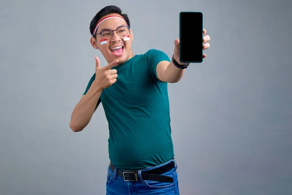 Excited young Asian man in casual t-shirt showing blank screen mobile phone, recommending mobile app isolated on grey background. indonesian independence day celebration concept