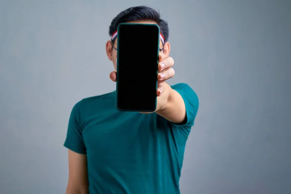 Handsome young Asian man in casual t-shirt showing mobile phone with blank screen, recommending mobile app isolated on grey background. indonesian independence day celebration concept