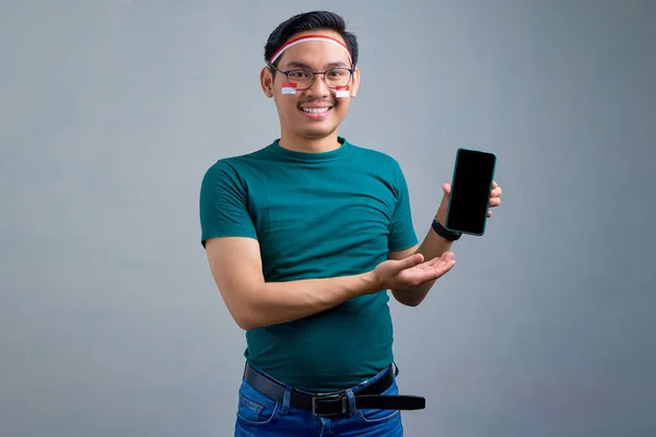 Cheerful young Asian man in casual t-shirt presenting mobile phone with blank screen, recommending mobile app isolated on grey background. indonesian independence day celebration concept