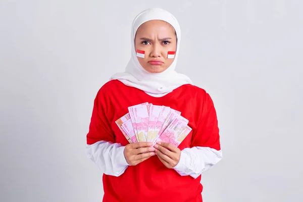 Confused young Asian muslim woman in red white t-shirt holding cash money in Indonesian rupiah banknotes isolated on white background. Indonesian independence day on 17 august concept
