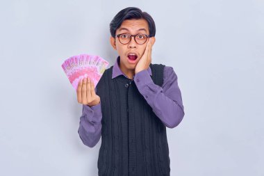 Gömlekli, yakışıklı, yakışıklı bir Asyalı adam ağzı açık yanağa dokunuyor, elinde rupiah banknotlarla nakit para tutuyor ve beyaz arka planda izole edilmiş kameraya bakıyor. Halk yaşam tarzı konsepti