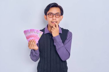 Gömlek giymiş, gülümseyen yakışıklı Asyalı bir adam tırnaklarını yiyor ve beyaz arka planda izole edilmiş rupiah banknotlarla nakit para tutuyor. Halk yaşam tarzı konsepti