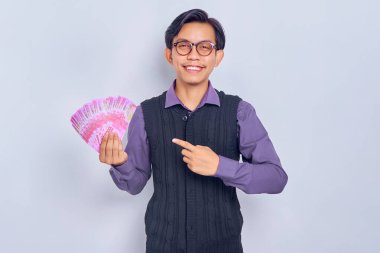 Gömlek giymiş, gülümseyen yakışıklı Asyalı bir adam beyaz arka planda izole edilmiş ellerde rupiah banknotlarıyla paralarını işaret ediyor. Halk yaşam tarzı konsepti