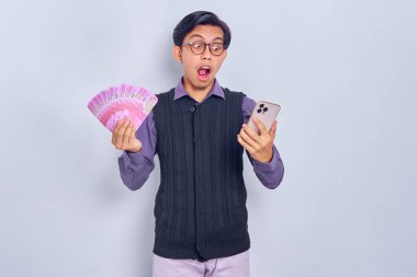 Mor gömlekli, cep telefonu kullanan ve beyaz arka planda izole edilmiş rupiah banknotlar halinde nakit para tutan Asyalı bir adamı şok etti. Halk yaşam tarzı konsepti
