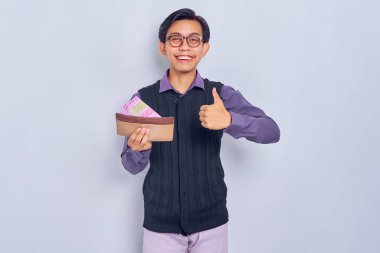 30 'lu yaşlarda gülümseyen Asyalı bir adamın portresi gömlek ve yelek giymiş kahverengi cüzdanı ve rupiah nakit para gösteriyor, beyaz arka planda izole edilmiş baş parmaklar gösteriyor.