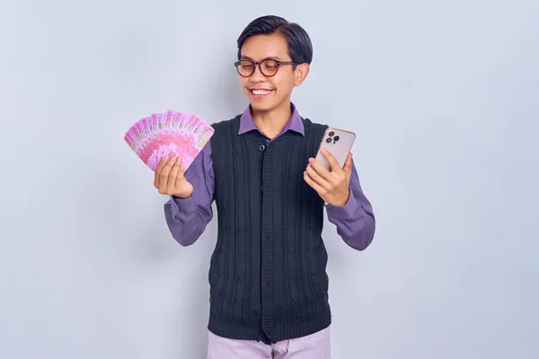 紫のシャツの服に身を包んだ若いアジア人男性が ルピア紙幣や携帯電話で現金を持ち 白い背景に隔離された笑顔 人々のライフスタイルコンセプト — ストック写真
