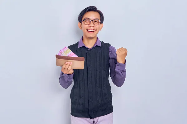 30代の興奮した若いアジア人男性の肖像シャツとベストを着て茶色の財布と現金お金でルピア 白の背景に隔離された大きな幸運を祝う — ストック写真