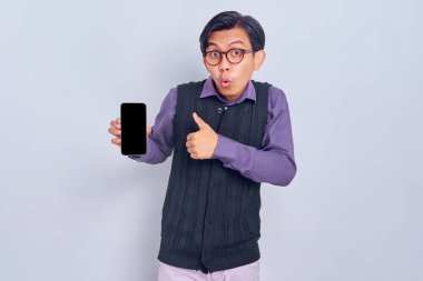 Boş ekranlı cep telefonu gösteren rahat gömlekli ve yelekli heyecanlı genç Asyalı adam, baş parmak hareketi yap, beyaz arka planda izole edilmiş mobil uygulama tavsiye et. Halk yaşam tarzı konsepti