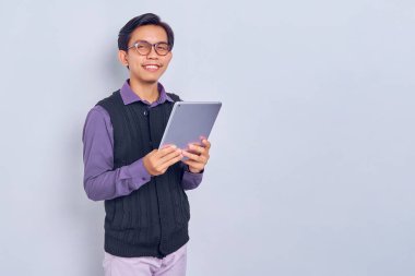 Beyaz arka planda izole edilmiş tablet bilgisayar kullanarak günlük gömlekli ve yelekli gülümseyen Asyalı genç adam. Halk yaşam tarzı konsepti