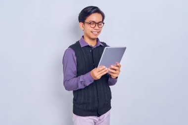 Beyaz arka planda izole edilmiş tablet bilgisayar kullanarak günlük gömlekli ve yelekli gülümseyen Asyalı genç adam. Halk yaşam tarzı konsepti