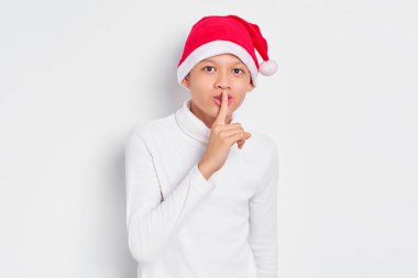 Beyaz tişörtlü, Noel Baba şapkalı sessiz jest yapan Asyalı bir çocuk beyaz arka planda sessiz kalmak istiyor. Mutlu Noeller ve mutlu yıllar.