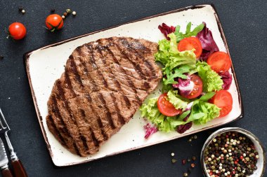 Salata ve sebzeli bir tabakta ızgara biftek ve koyu bir arka plan. Akşam yemeği.
