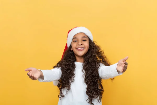 提起一个黑皮肤 留着长长的卷发 头戴圣诞老人帽的小女孩的画像 祝贺圣诞节快乐 她在黄色背景下向前伸出双手 — 图库照片