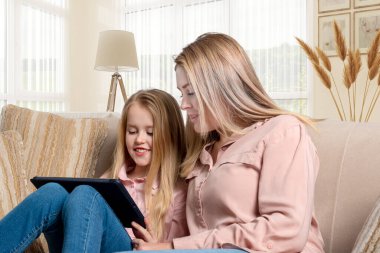 Uzun saçlı, pembe tişörtlü ve kot pantolonlu bir anne ve kızının evdeki kanepede dizüstü bilgisayarla oturması..  