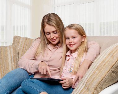 Uzun saçlı, pembe tişörtlü ve kot pantolonlu bir anne ve kızının evdeki kanepede dizüstü bilgisayarla oturması..  