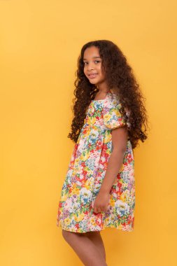 Half length portrait of a dark skinned young girl with long curly hair wearing colorfull dress against yellow background