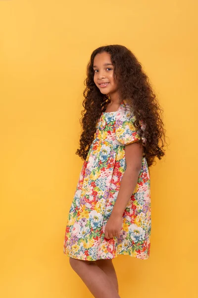 Half length portrait of a dark skinned young girl with long curly hair wearing colorfull dress against yellow background