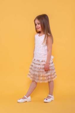 Full length portrait of a  young  smiling girl with long blonde hair wearing white dress against yellow background  in studio