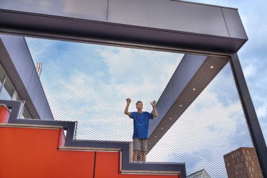 Shot of a young teen boy in a blue T-shirt standing upset about something on a red ladder in the street