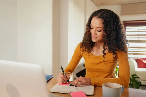 Feliz Mujer multicultural enfocada, escribiendo en cuaderno. Joven profesional trabajando a distancia con el ordenador portátil en apartamento moderno — Foto de Stock