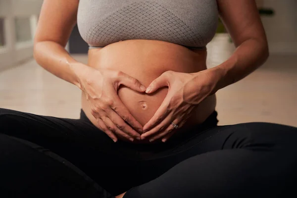 Kafkasyalı hamile anne yoga minderinde oturuyor ve elleri karnında kalp şekli oluşturuyor.