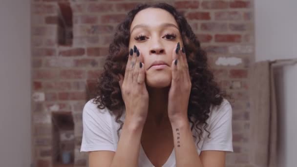 Multi-culturele jonge vrouw wrijven in het gezicht moisturizer terwijl glimlachen in de spiegel in de moderne badkamer — Stockvideo
