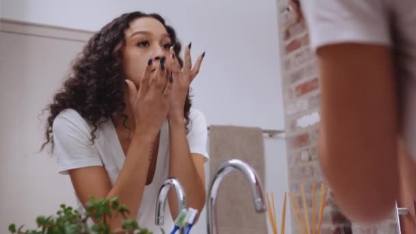 Πολυπολιτισμική νεαρή γυναίκα τρίβει σε ενυδατική κρέμα στο πρόσωπό της στο σύγχρονο καθρέφτη του μπάνιου. Χαμογελαστό, υγιές δέρμα — Αρχείο Βίντεο