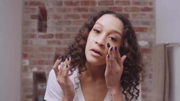 Multi-culturele jonge vrouw aanbrengen gezicht moisturizer terwijl glimlachen in de spiegel in de moderne badkamer — Stockvideo