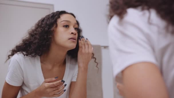 Multi-culturele jonge vrouw op zoek naar vlekken op haar gezicht in moderne badkamer spiegel — Stockvideo