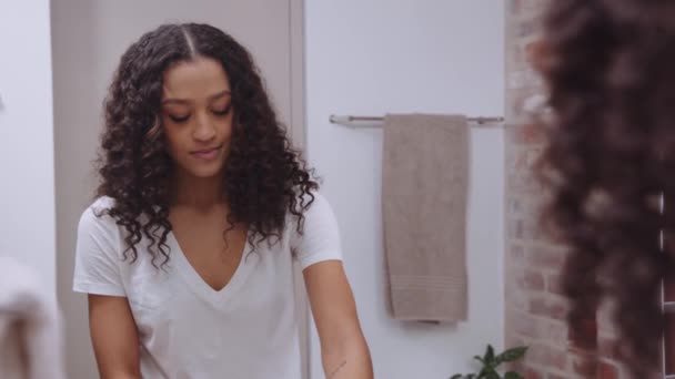 Молодая афроамериканка улыбается, чистя зубы перед сном, глядя в зеркало в современной ванной комнате — стоковое видео