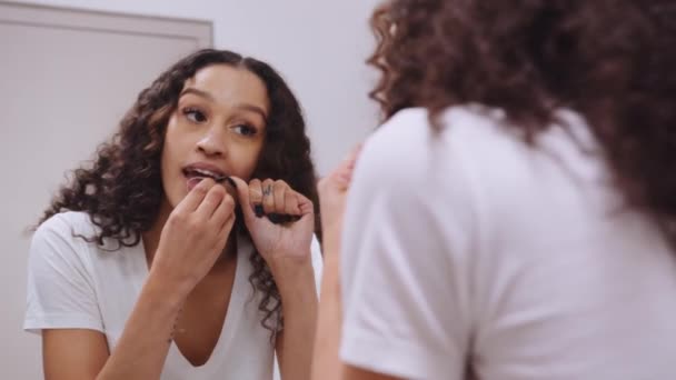 Afro-américaine jeune femelle fil dentaire ses dents avec fil dentaire dans le miroir dans sa salle de bain moderne — Video