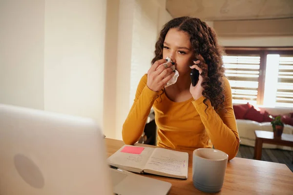 Joven mujer multicultural sonándose la nariz mientras hace una llamada de negocios. Enfermo y trabajando desde casa en el ordenador portátil — Foto de Stock
