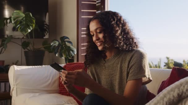 Multiculturele jonge volwassen vrouw sms 'en vrienden terwijl ze op de bank zit te glimlachen. Modern appartement, vrijetijdsbesteding overdag — Stockvideo