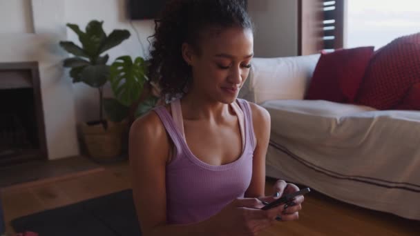 Felice Multi-culturale giovane donna adulta ridendo e digitando sul suo telefono mentre sul tappetino yoga in salotto moderno a casa — Video Stock